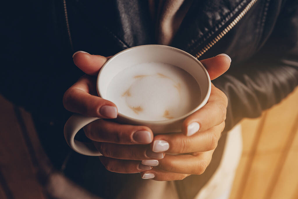 close up fêmea vestida com roupas de outono segura em duas mãos uma grande xícara de café quente com leite em pé na rua. A menina aquece a xícara e gosta de café aromático
 - Foto, Imagem