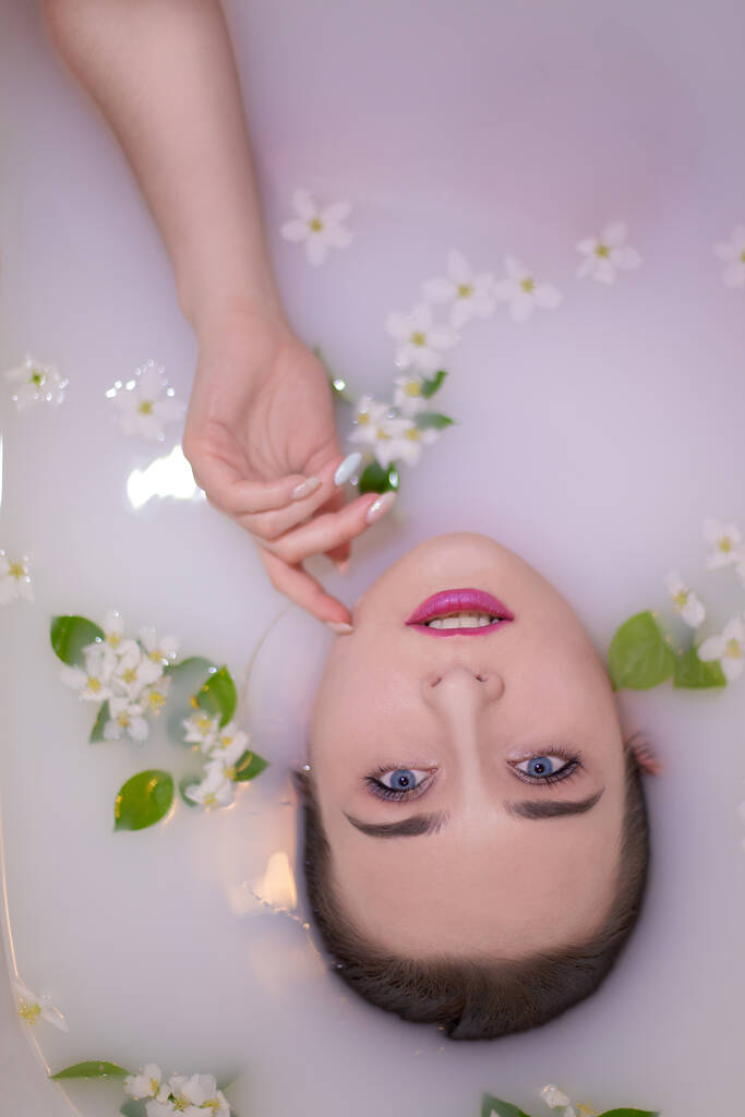 Urocza dziewczyna ze stylowym makijażem i pięknymi niebieskimi oczami bierze kąpiel w mleku z różowym odcieniem rozrzuconym kwiatami i liśćmi jabłka. Dotyka słodkiej twarzy delikatnymi dłońmi. - Zdjęcie, obraz