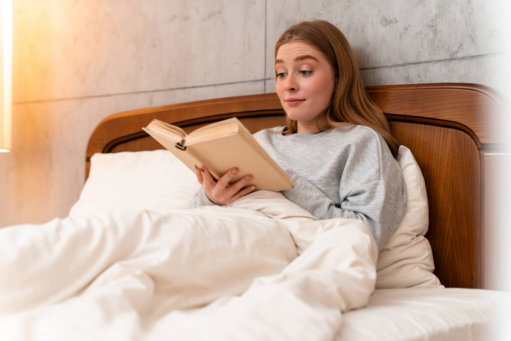 Φωτογραφία από μια έκπληκτη όμορφη νεαρή γυναίκα σε εσωτερικούς χώρους στο σπίτι στο κρεβάτι διαβάζοντας το βιβλίο. - Φωτογραφία, εικόνα