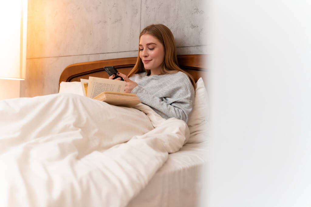Fénykép egy pozitív optimista csinos fiatal nő beltéri otthon az ágyban könyv segítségével mobiltelefon. - Fotó, kép