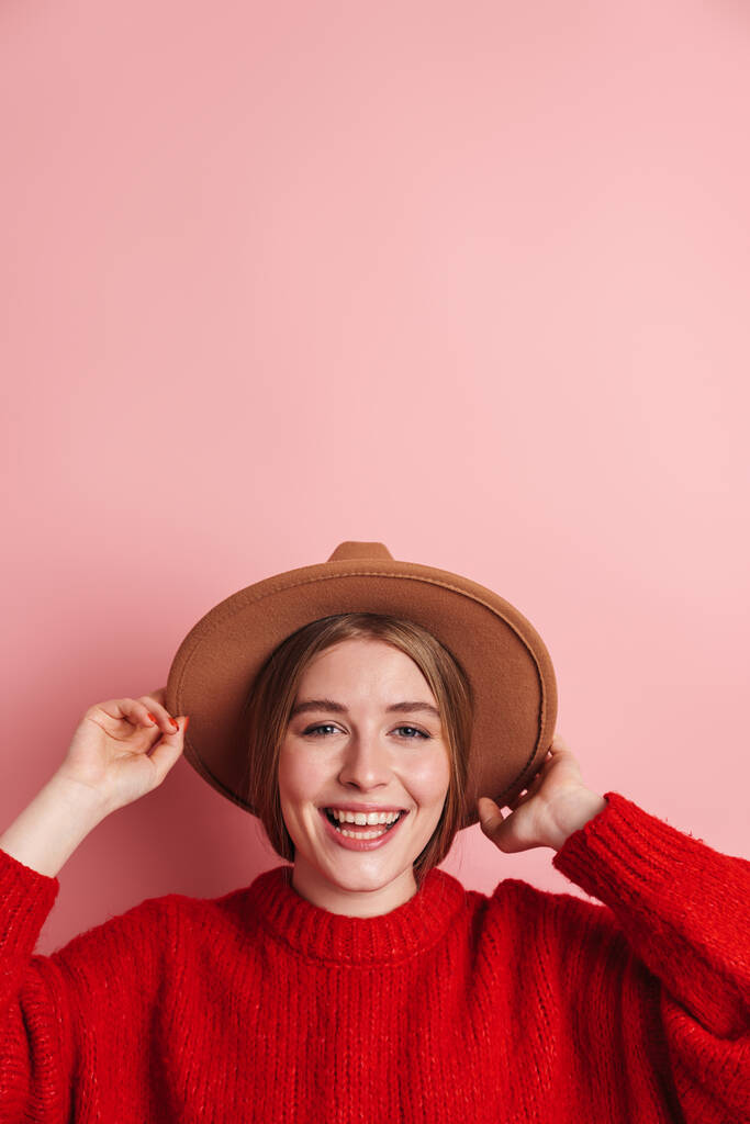 Foto de mulher bonita feliz em camisola vermelha sorrindo e segurando chapéu isolado sobre a parede rosa
 - Foto, Imagem