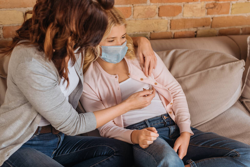 Mulher segurando termômetro perto da filha em máscara médica no sofá
  - Foto, Imagem
