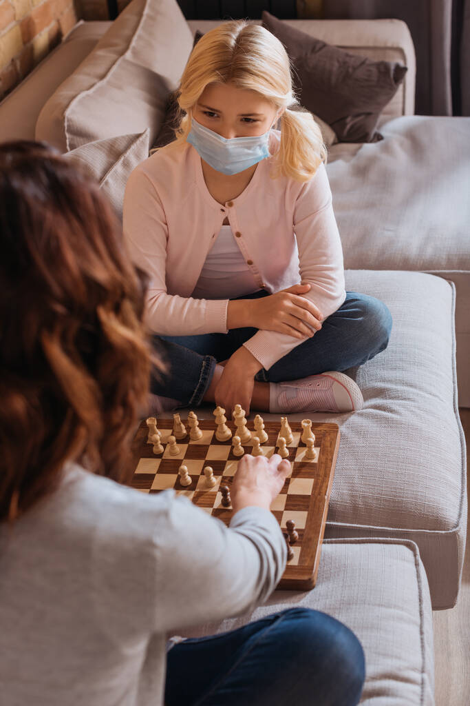 Tıbbi maskeli bir çocuğun annesiyle kanepede satranç oynayacağı seçmeli bir odak noktası.  - Fotoğraf, Görsel