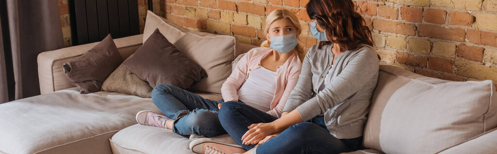 Orientação panorâmica de mãe e filha em máscaras médicas sentadas no sofá em casa
  - Foto, Imagem