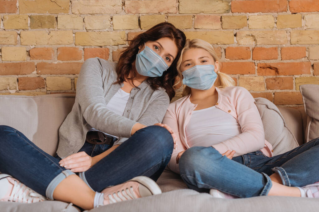 Mother and kid in medical masks looking at camera on couch at home  - Photo, Image