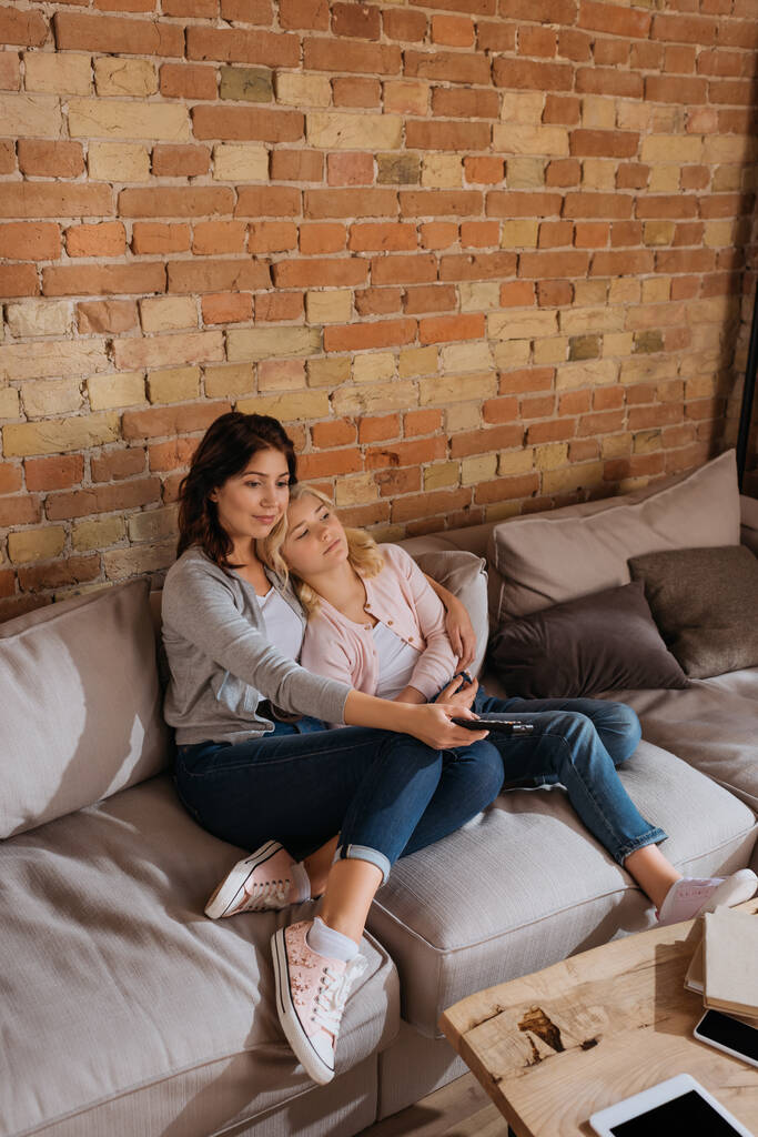 Anya átöleli gyermek, miközben tartja távirányító a kanapén  - Fotó, kép