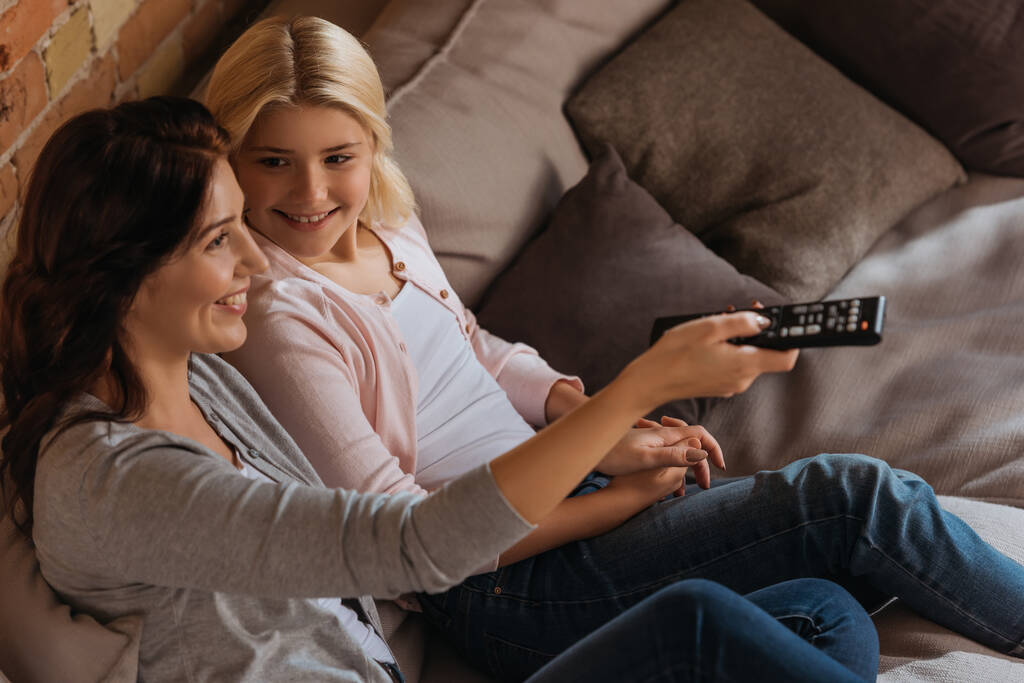 Selektivní zaměření usměvavé dítě při pohledu na matku s dálkovým ovladačem v obývacím pokoji - Fotografie, Obrázek