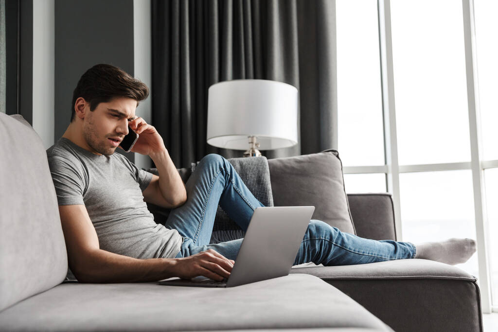 Porträt eines attraktiven ernsten jungen bärtigen Mannes in lässiger Kleidung, der auf einer Couch im Wohnzimmer sitzt und mit dem Laptop telefoniert - Foto, Bild