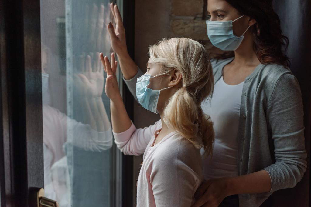 Selective focus of mother and daughter in medical masks standing near window at home  - Photo, Image