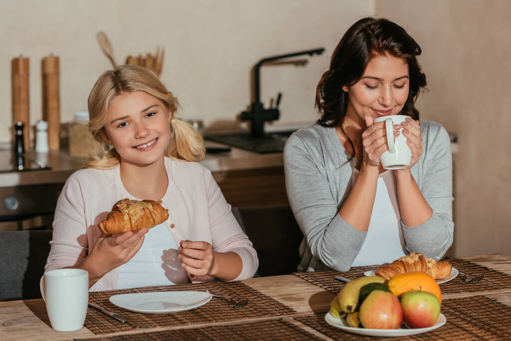 Lächelndes Kind blickt in die Kamera, während es in der Küche Croissant neben Mutter mit Tasse hält  - Foto, Bild
