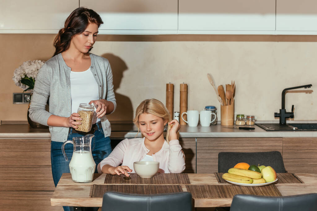 Mosolygós gyerek ül az asztalnál, közel anya gazdaság üveg gabonafélék a konyhában  - Fotó, kép