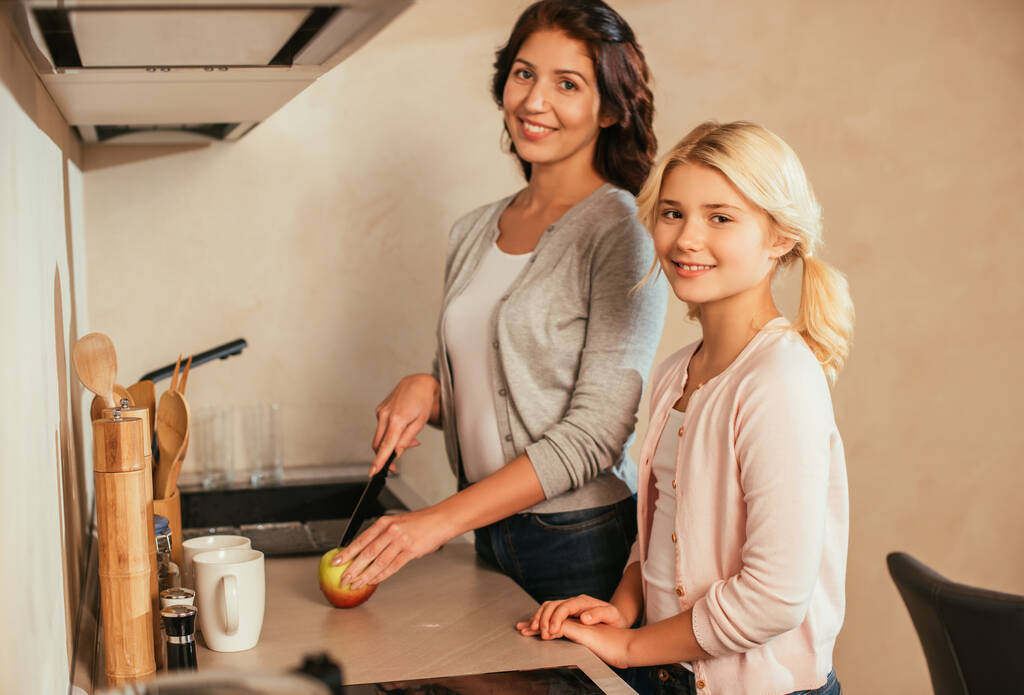 Vista lateral de mãe e filha sorridente olhando para a câmera enquanto corta maçã na bancada da cozinha
  - Foto, Imagem