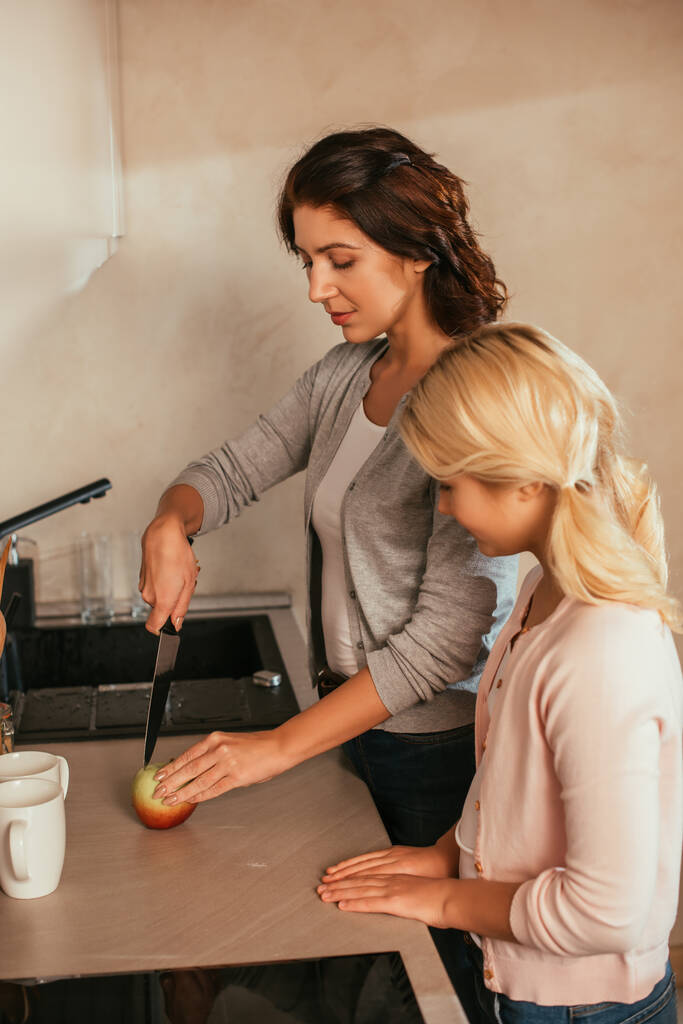 Πλευρική άποψη της κόρης κοιτάζοντας τη μητέρα κοπής μήλο στον πάγκο της κουζίνας  - Φωτογραφία, εικόνα