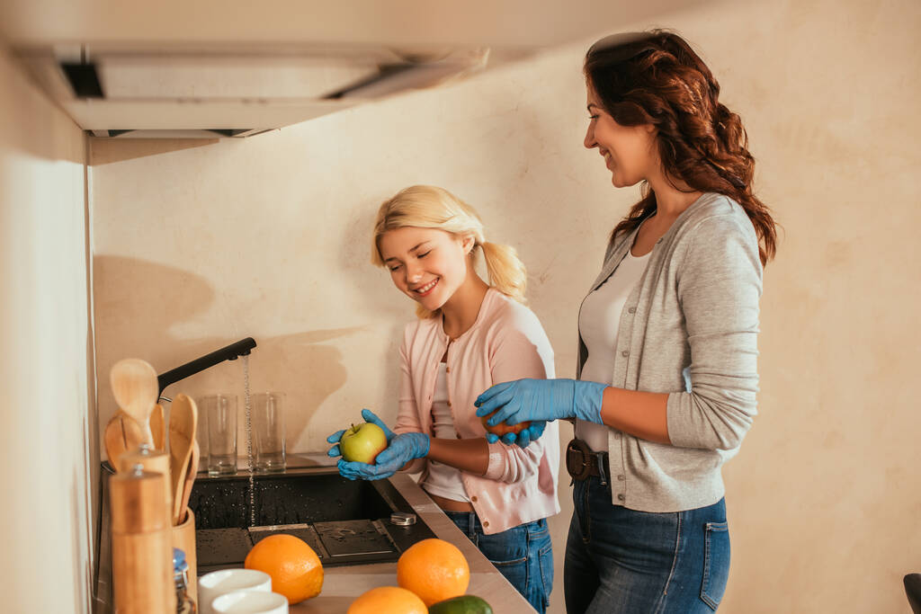 Селективный фокус улыбающейся матери и ребенка в латексных перчатках, моющих фрукты на кухне
  - Фото, изображение