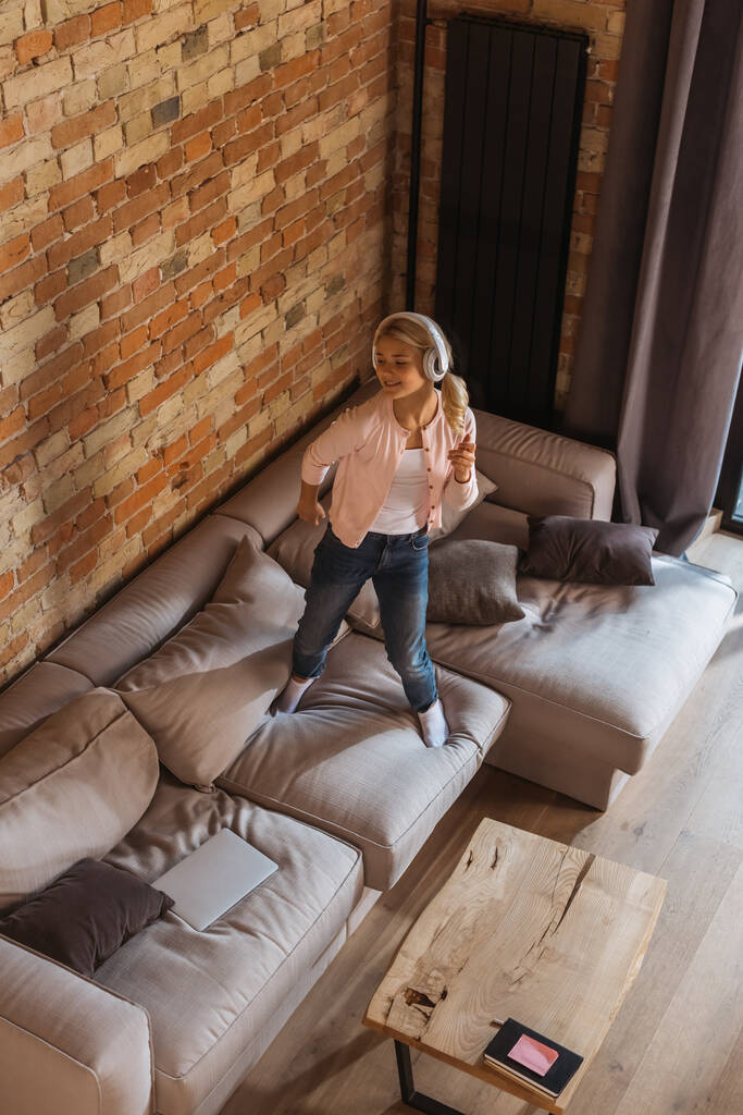 High angle view of cheerful child in headphones standing on couch near laptop at home - Photo, Image