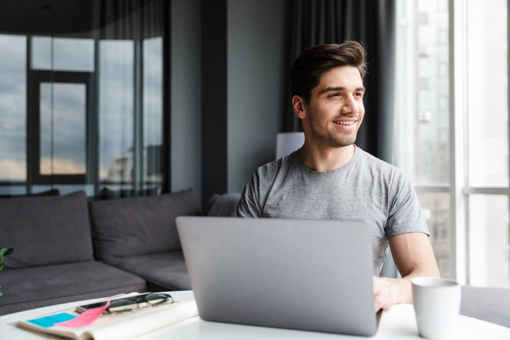 Jóképű, mosolygós, fiatal, szakállas férfi alkalmi ruhát visel, laptoppal, miközben otthon ül az asztalnál. - Fotó, kép