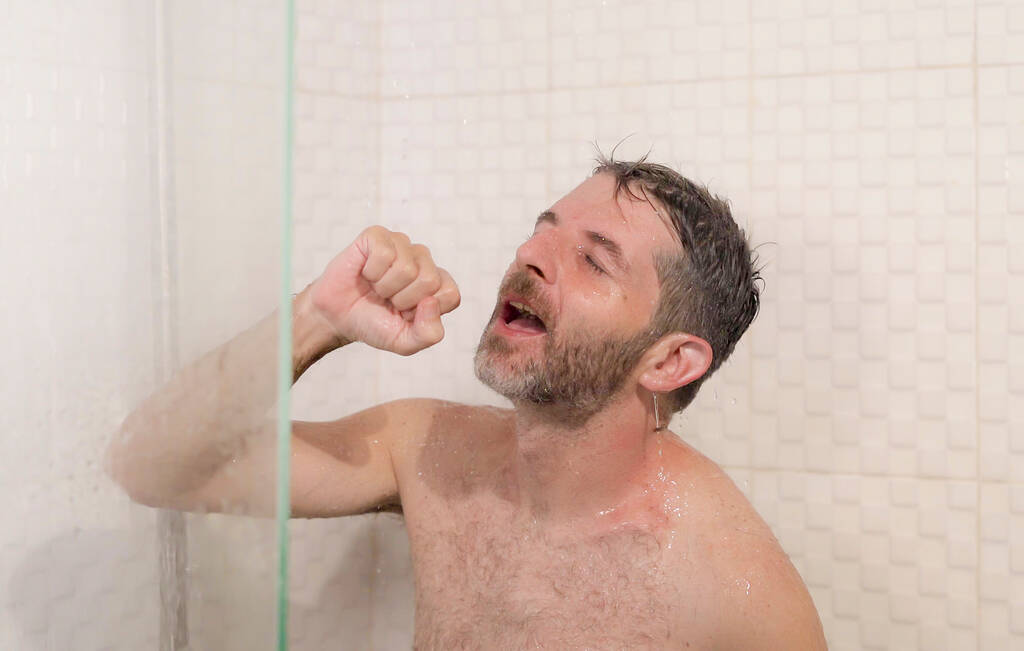 Lustiges Porträt eines jungen attraktiven und glücklichen Mannes mit Bart, der sorglos und verrückt zu Hause unter der Dusche singt und vorgibt, ein Mikrofon in der Hand zu halten und die Morgendusche genießt - Foto, Bild