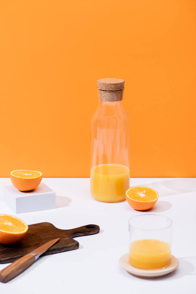 świeży sok pomarańczowy w szklance i butelce w pobliżu ciętych owoców na drewnianej desce do krojenia z nożem na białej powierzchni odizolowanej na pomarańczowo - Zdjęcie, obraz