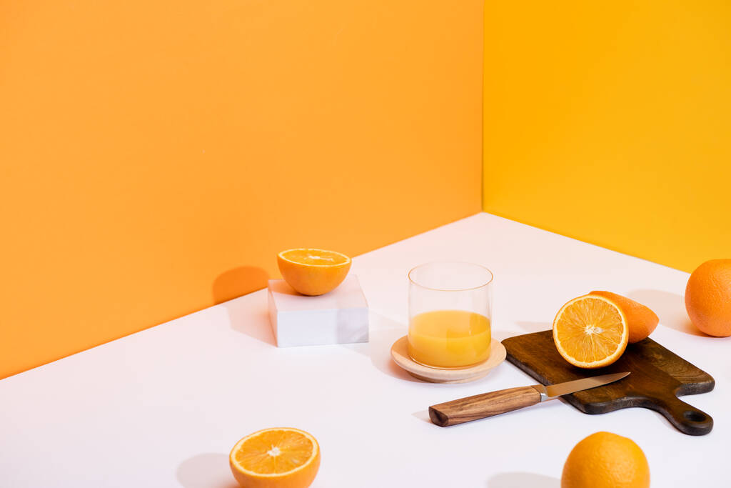 suco de laranja fresco em vidro perto de laranjas maduras, placa de corte de madeira com faca na superfície branca no fundo laranja
 - Foto, Imagem