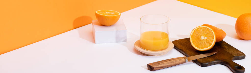 frischer Orangensaft im Glas bei reifen Orangen, Holzschneidebrett mit Messer auf weißer Oberfläche auf orangefarbenem Hintergrund, Panoramaaufnahme - Foto, Bild