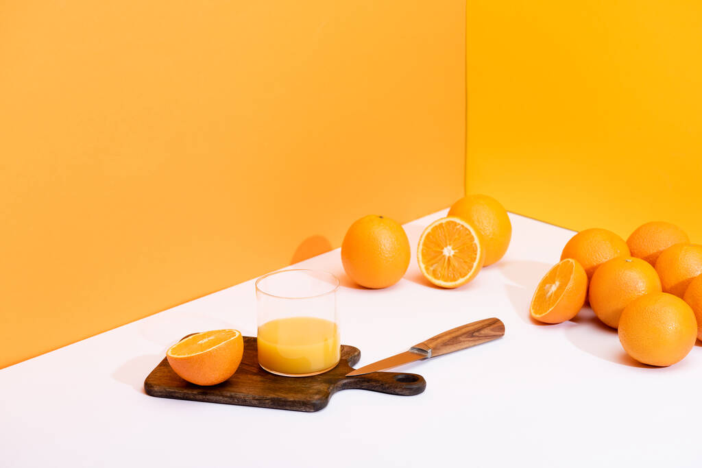 čerstvý pomerančový džus ve skle na dřevěné řezací desce s nožem v blízkosti zralých pomerančů na bílém povrchu na oranžovém pozadí - Fotografie, Obrázek