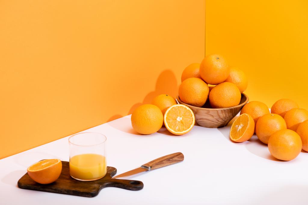 frischer Orangensaft im Glas auf Holzschneidebrett mit Messer in der Nähe reifer Orangen in Schüssel auf weißer Oberfläche auf orangefarbenem Hintergrund - Foto, Bild