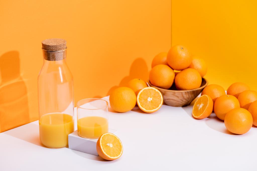 φρέσκος χυμός πορτοκαλιού σε ποτήρι και μπουκάλι κοντά σε ώριμα πορτοκάλια σε μπολ σε λευκή επιφάνεια σε πορτοκαλί φόντο - Φωτογραφία, εικόνα