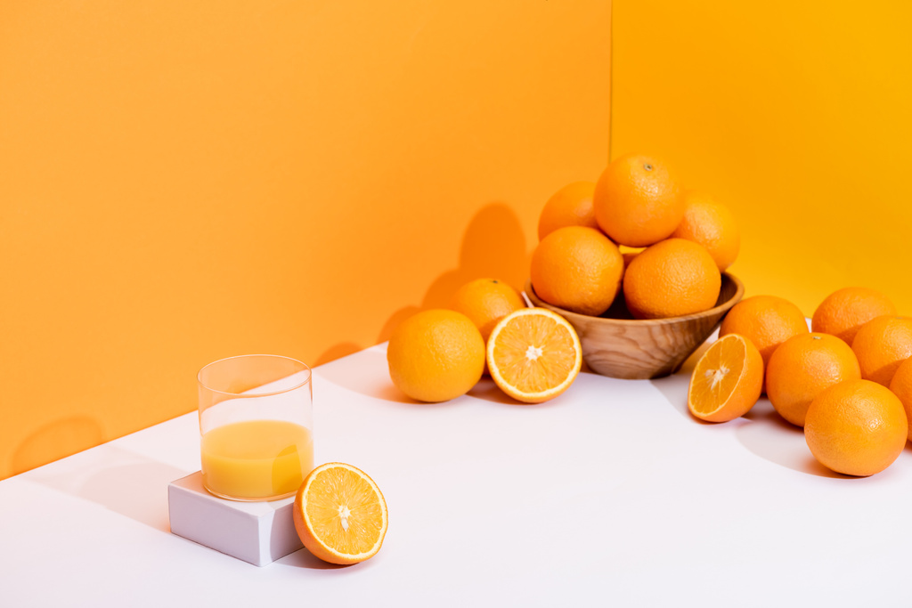 świeży sok pomarańczowy w szkle w pobliżu dojrzałych pomarańczy w misce na białej powierzchni na pomarańczowym tle - Zdjęcie, obraz