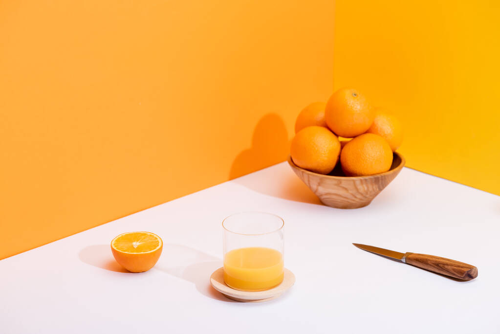 frischer Orangensaft im Glas neben reifen Orangen in Schüssel und Messer auf weißer Oberfläche auf orangefarbenem Hintergrund - Foto, Bild