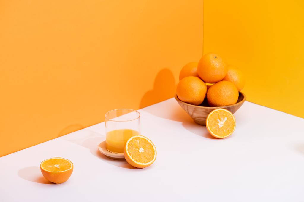 čerstvý pomerančový džus ve skle u zralých pomerančů v misce na bílém povrchu na oranžovém pozadí - Fotografie, Obrázek