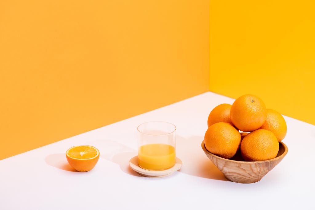 frischer Orangensaft im Glas neben reifen Orangen in Schüssel auf weißer Oberfläche auf orangefarbenem Hintergrund - Foto, Bild