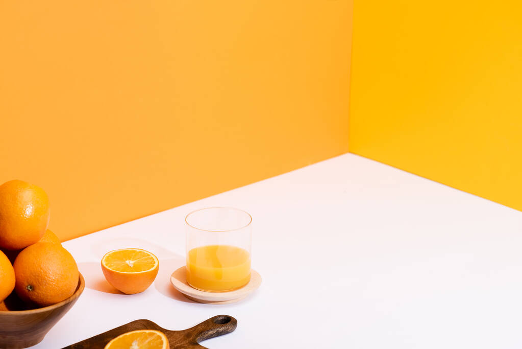 zumo de naranja fresco en vaso cerca de naranjas maduras en tazón y tabla de cortar de madera sobre superficie blanca sobre fondo naranja
 - Foto, Imagen