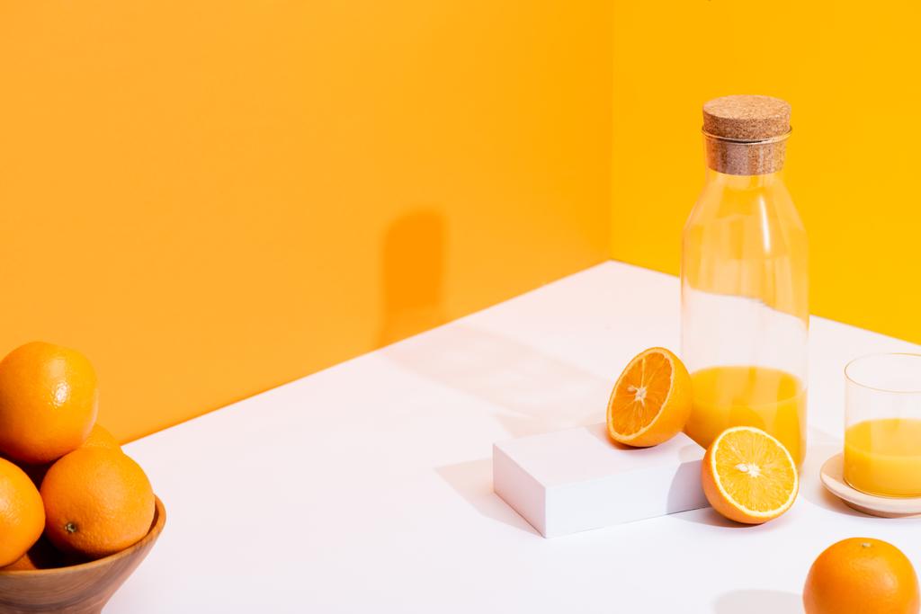 świeży sok pomarańczowy w szklanej butelce w pobliżu dojrzałych pomarańczy w misce na białej powierzchni na pomarańczowym tle - Zdjęcie, obraz