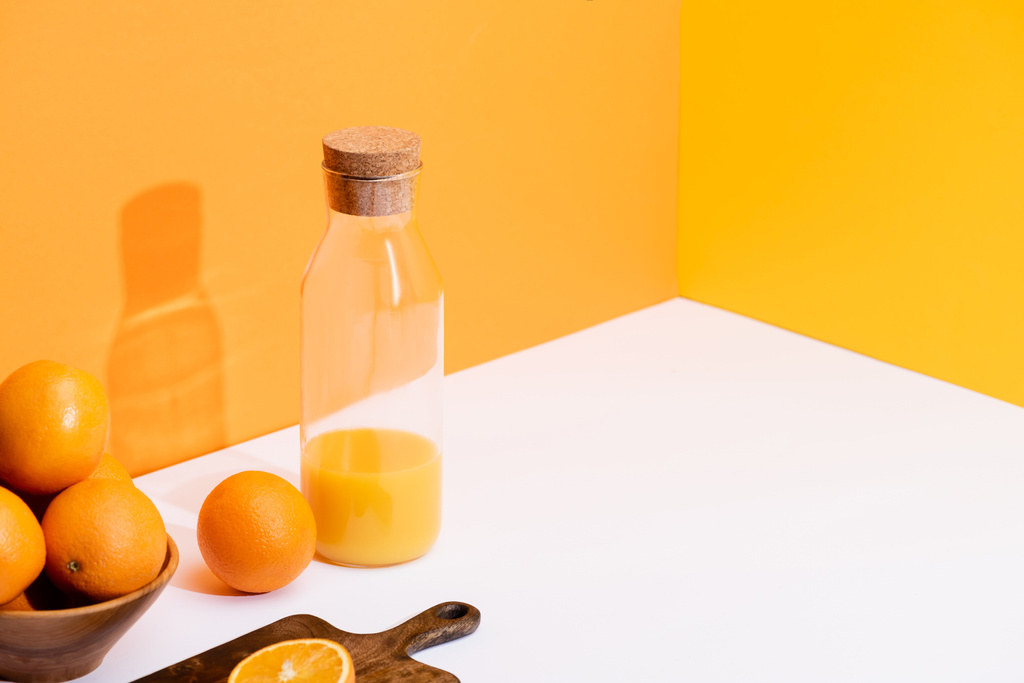 φρέσκος χυμός πορτοκαλιού σε γυάλινο μπουκάλι κοντά σε ώριμα πορτοκάλια σε μπολ και ξύλο κοπής σε λευκή επιφάνεια σε πορτοκαλί φόντο - Φωτογραφία, εικόνα