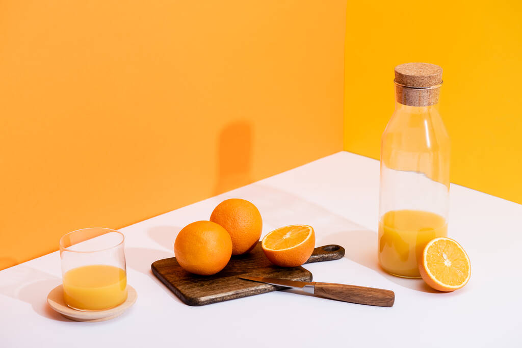 vers sinaasappelsap in glas en fles in de buurt van rijpe sinaasappels op snijplank met mes op wit oppervlak op oranje achtergrond - Foto, afbeelding