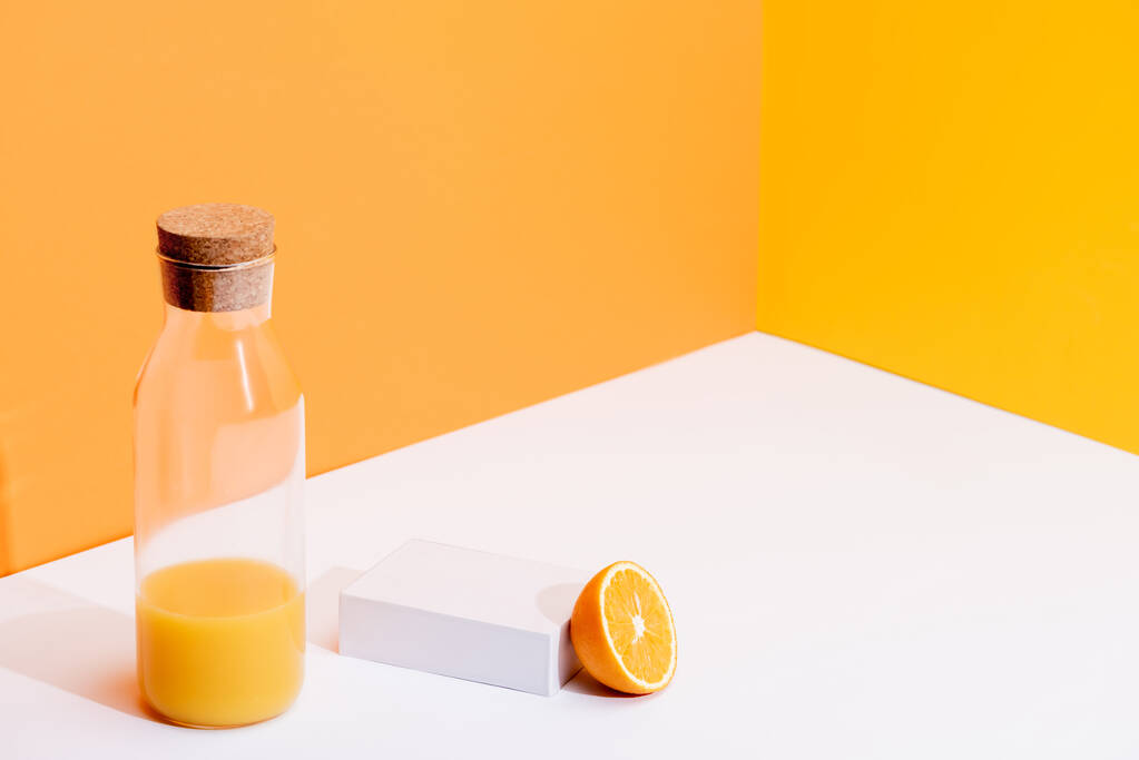 čerstvý pomerančový džus ve skleněné láhvi blízko zralého pomeranče na bílém povrchu na oranžovém pozadí - Fotografie, Obrázek