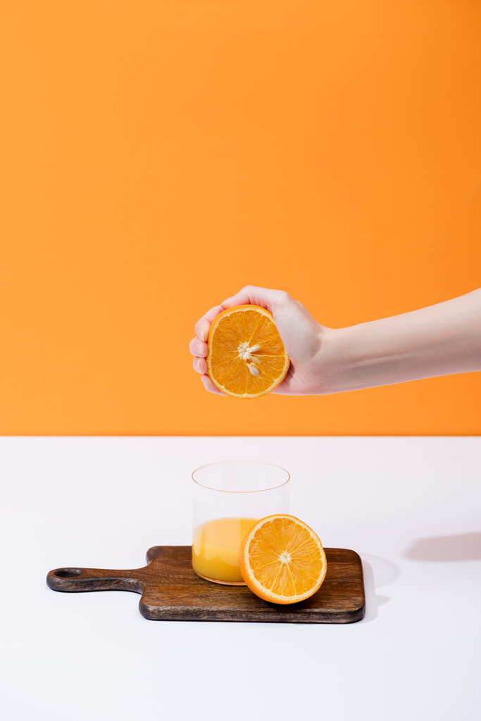 oříznutý pohled ženy mačkající čerstvý pomerančový džus ve skle na dřevěné řezací desce na bílém povrchu izolovaném na oranžové - Fotografie, Obrázek