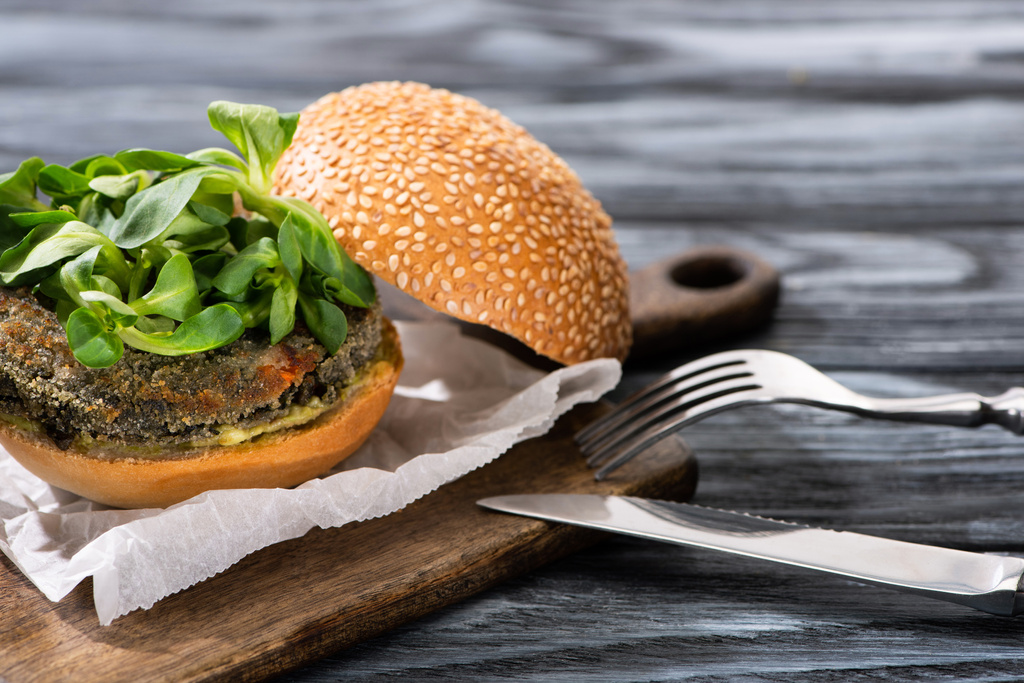 smakelijke veganistische hamburger met microgreens geserveerd op snijplank met bestek op houten tafel - Foto, afbeelding