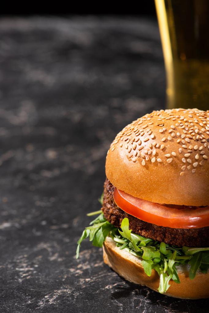selectieve focus van smakelijke veganistische hamburger met tomaat en groenten geserveerd op getextureerd oppervlak met bier - Foto, afbeelding