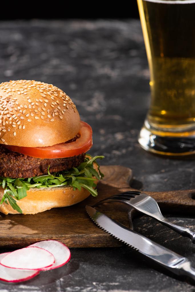 selektiver Schwerpunkt des leckeren veganen Burgers mit Tomate und Gemüse, serviert auf Holzbrett auf strukturierter Oberfläche mit Bier und Besteck - Foto, Bild