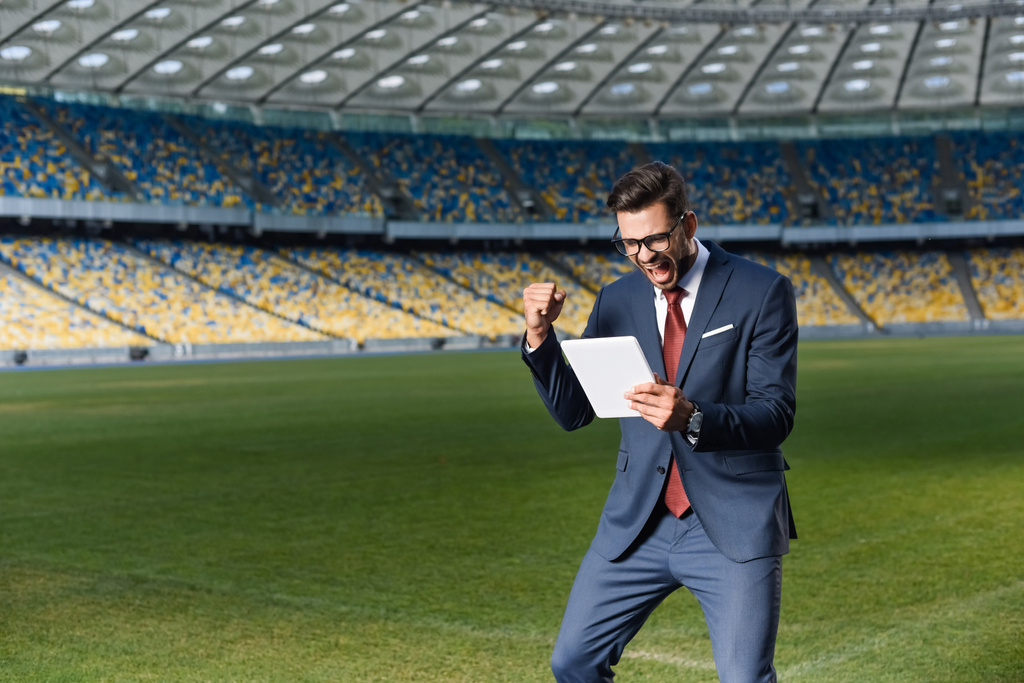 young businessman in suit and glasses using digital tablet and showing yeas gesture at stadium, sports betting concept - Photo, Image