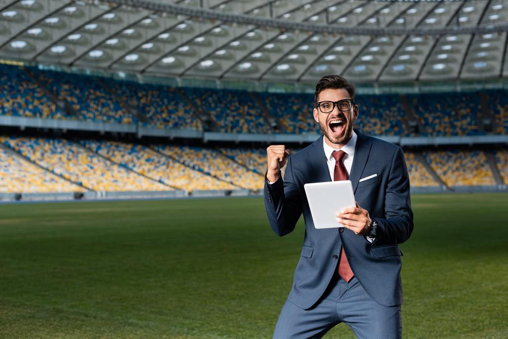 young businessman in suit and glasses using digital tablet and showing yeas gesture at stadium, sports betting concept - Photo, Image