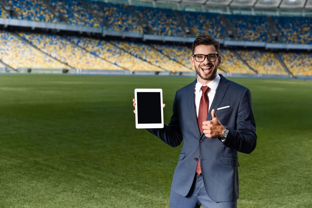 onnellinen nuori liikemies puku ja lasit tilalla digitaalinen tabletti tyhjällä näytöllä ja näyttää peukalo ylös stadionilla
 - Valokuva, kuva