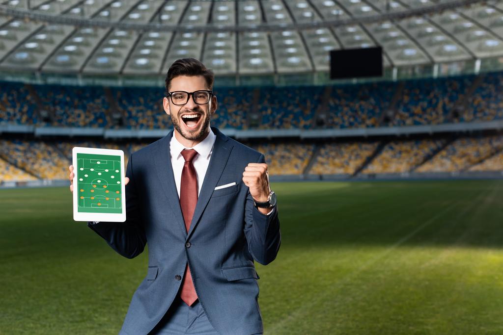 veselý mladý podnikatel v obleku a brýle drží digitální tablet s fotbalovou hru na obrazovce a ukazuje ano gesto na stadionu - Fotografie, Obrázek