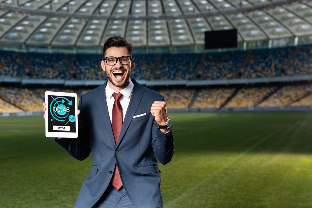 vrolijke jonge zakenman in pak en bril met digitale tablet met app en met ja gebaar in het stadion - Foto, afbeelding