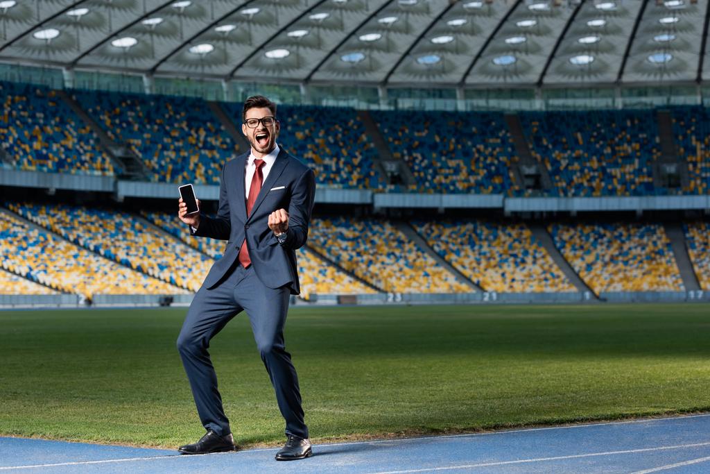 full length view of excited young businessman in suit and glasses holding smartphone with blank screen and showing yes gesture at stadium - Photo, Image