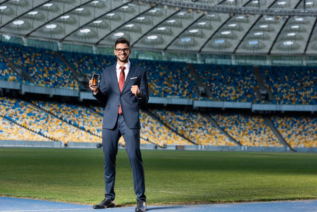 usmívající se mladý podnikatel v obleku a brýle drží smartphone s grafy a grafy na obrazovce a ukazuje ano gesto na stadionu - Fotografie, Obrázek