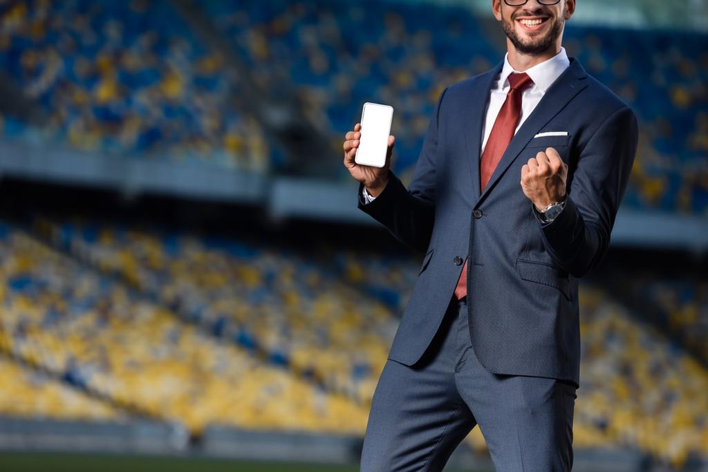 Ausgeschnittene Ansicht eines lächelnden jungen Geschäftsmannes im Anzug, der sein Smartphone mit leerem Bildschirm hält und im Stadion Ja-Geste zeigt - Foto, Bild
