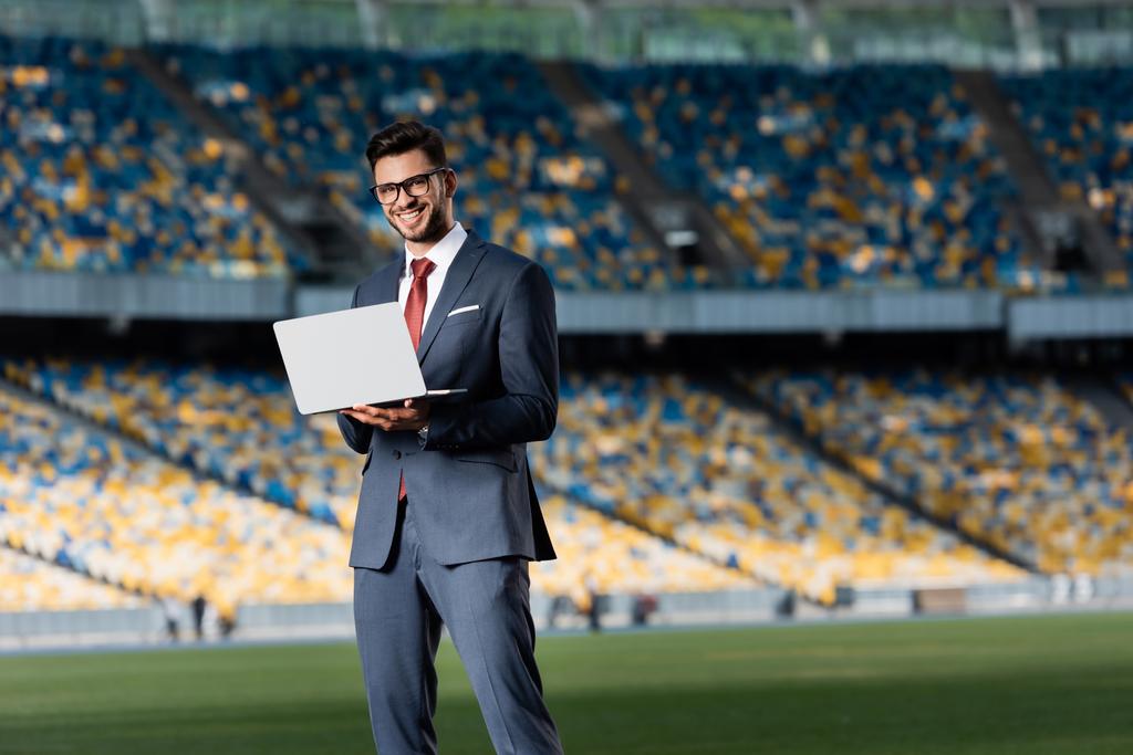 smiling young businessman in suit with laptop at stadium - Photo, Image