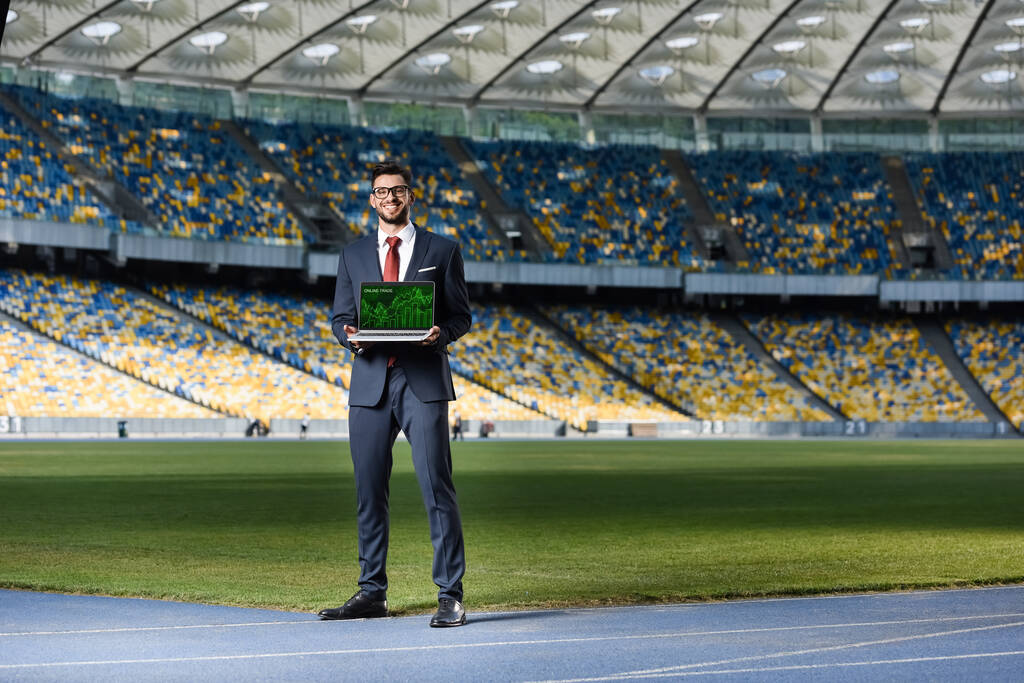 uśmiechnięty młody biznesmen w garniturze pokazujący laptopa ze stroną internetową handlu online na stadionie - Zdjęcie, obraz